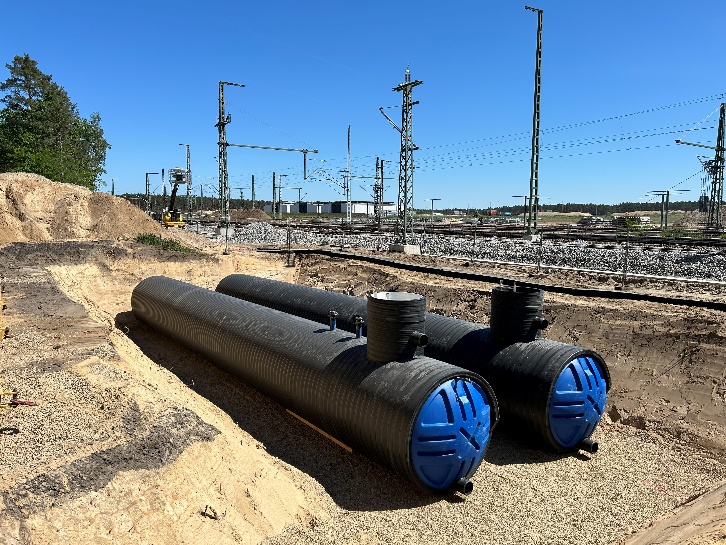 Zwei schwarze HDPE-Löschwassertanks mit blauen Endböden und aufgesetzten Domschächten liegen in einer Sandbaugrube, dahinter ein Gleisfeld mit Oberleitungsmasten.